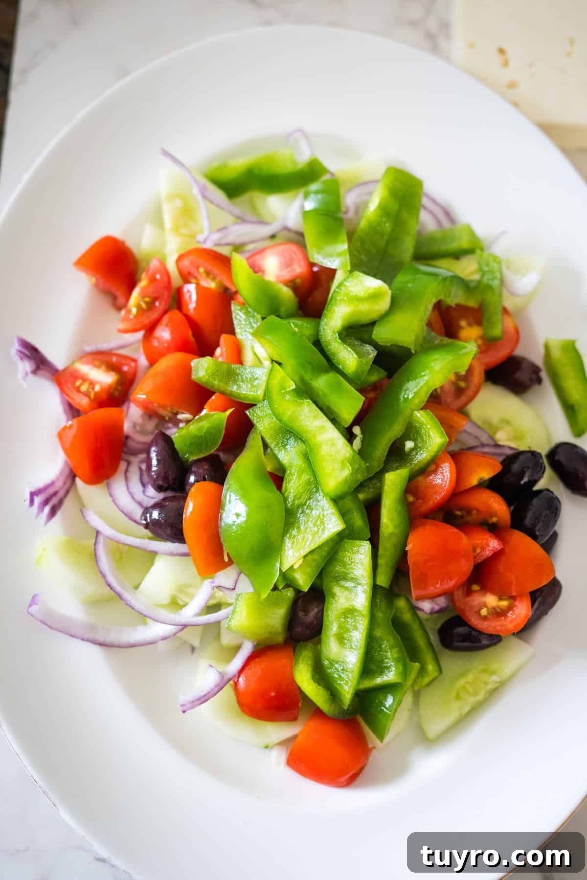 Authentic Greek Horiatiki Salad Recipe — Classic Village Salad 7 A white plate with a Greek village salad of sliced green bell peppers, red cherry tomatoes, black olives, red onion, and cucumber slices.