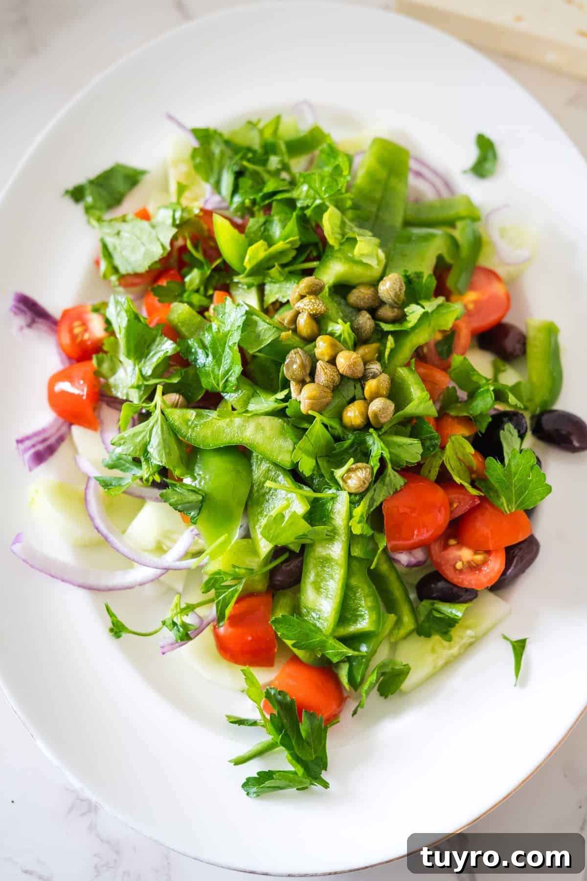 Authentic Greek Horiatiki Salad Recipe — Classic Village Salad 8 A Greek village salad with cherry tomatoes, snow peas, red onion, parsley, capers, and olives on a white plate.