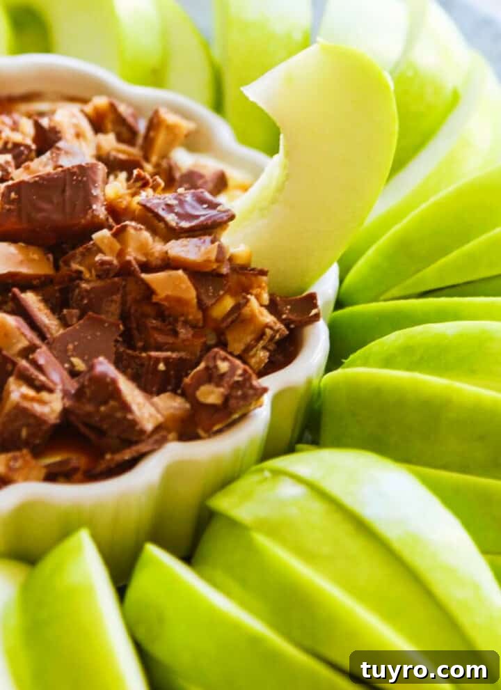 Slices of green Granny Smith apples around a white dish with a sweet dessert dip inside a low small white dish with a ruffled edge, and a piece of apple sticking into the dip.
