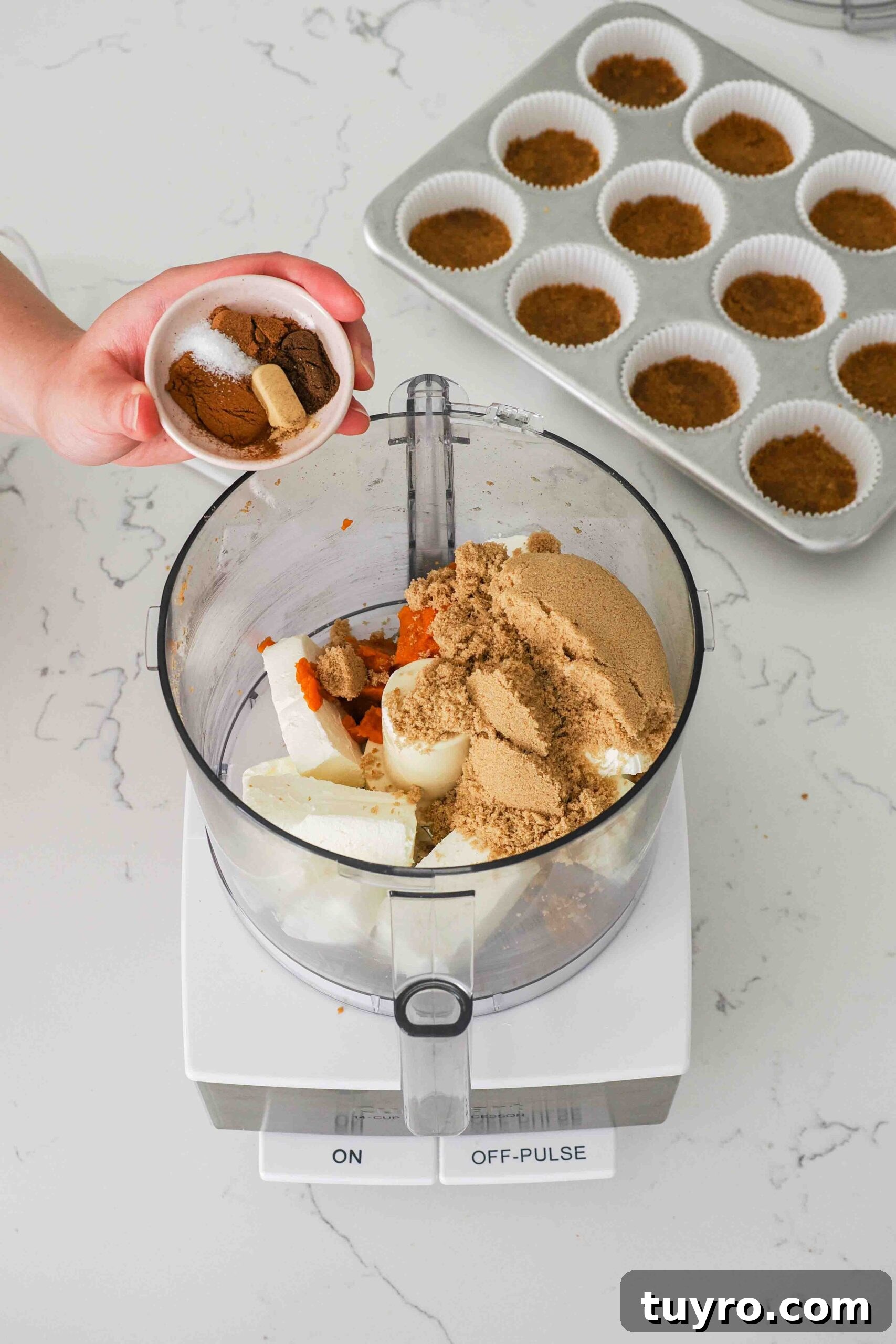 Mini Pumpkin Cheesecake Bites with Spiced Gingersnap Crust 7 A hand adds spices into a food processor bowl with cream cheese, pumpkin, and brown sugar.