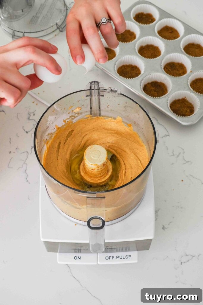 Mini Pumpkin Cheesecake Bites with Spiced Gingersnap Crust 9 Two hands crack an egg into a food processor bowl with the pumpkin cheesecake.