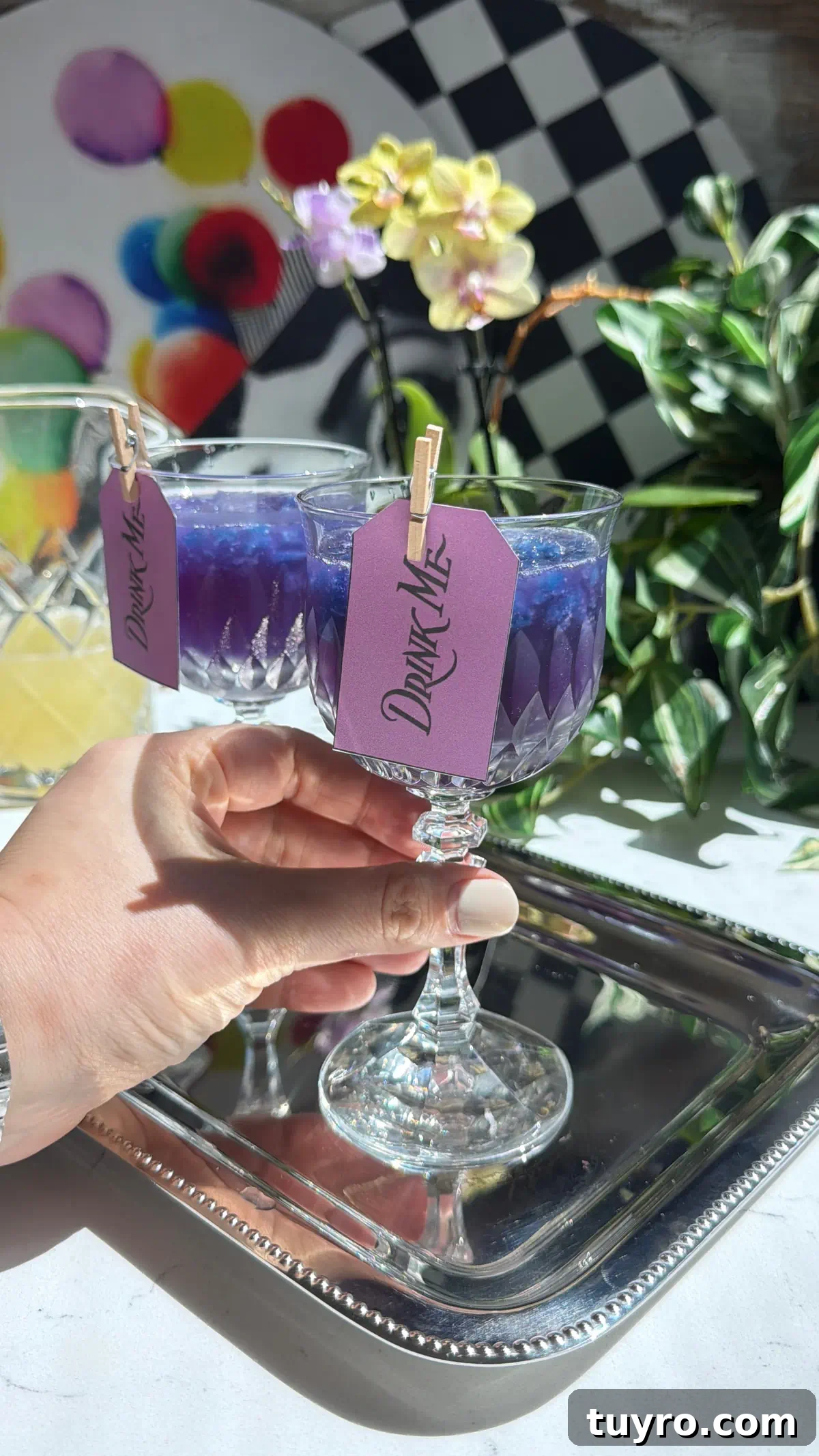Galaxy Cotton Candy Lemonade Cocktail — Color-Changing Drink Tutorial 5 A hand holding a crystal glass filled with a purple color-changing galaxy cocktail over shaved blue butterfly pea granita, featuring a Drink Me tag.