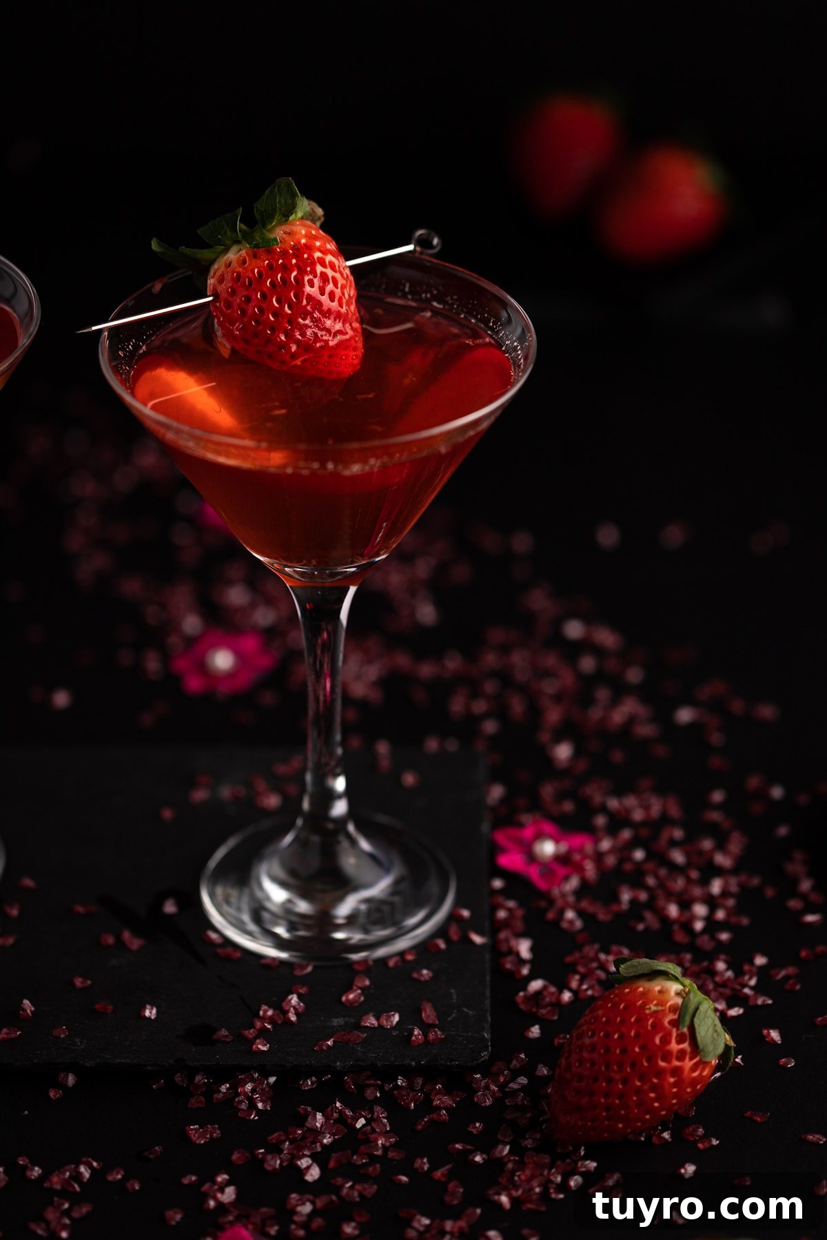 Strawberry martini, garnished with a strawberry, on a black background, with pink pebbles and flowers scattered around.