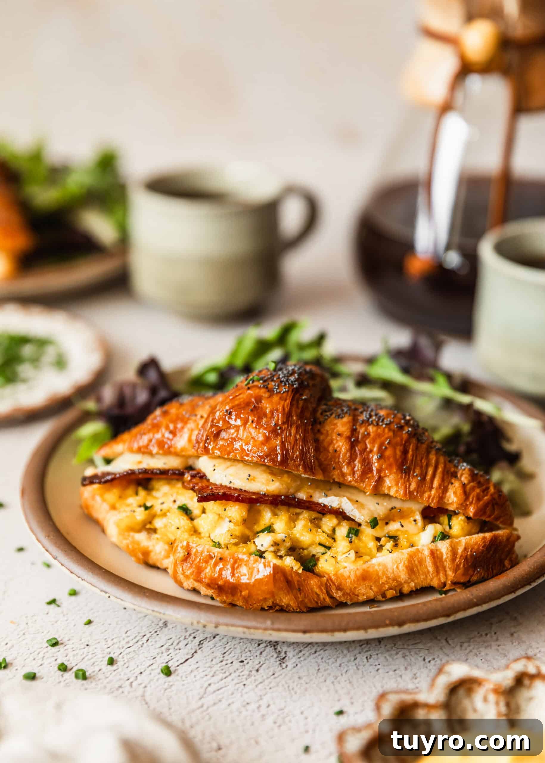 Sheet Pan Bacon, Egg & Cheese Croissant Sandwiches for Brunch 2 A bacon, egg, and cheese croissant sandwich next to greens on a tan plate with coffee and a bowl of Dijon sauce.