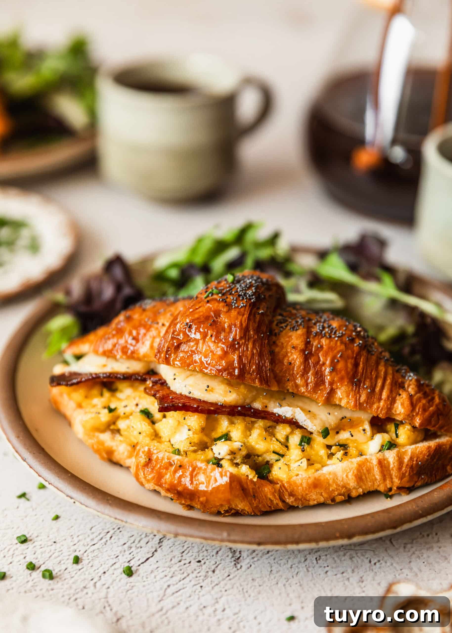 Sheet Pan Bacon, Egg & Cheese Croissant Sandwiches for Brunch 10 A croissant breakfast sandwich on a plate next to cups of coffee.