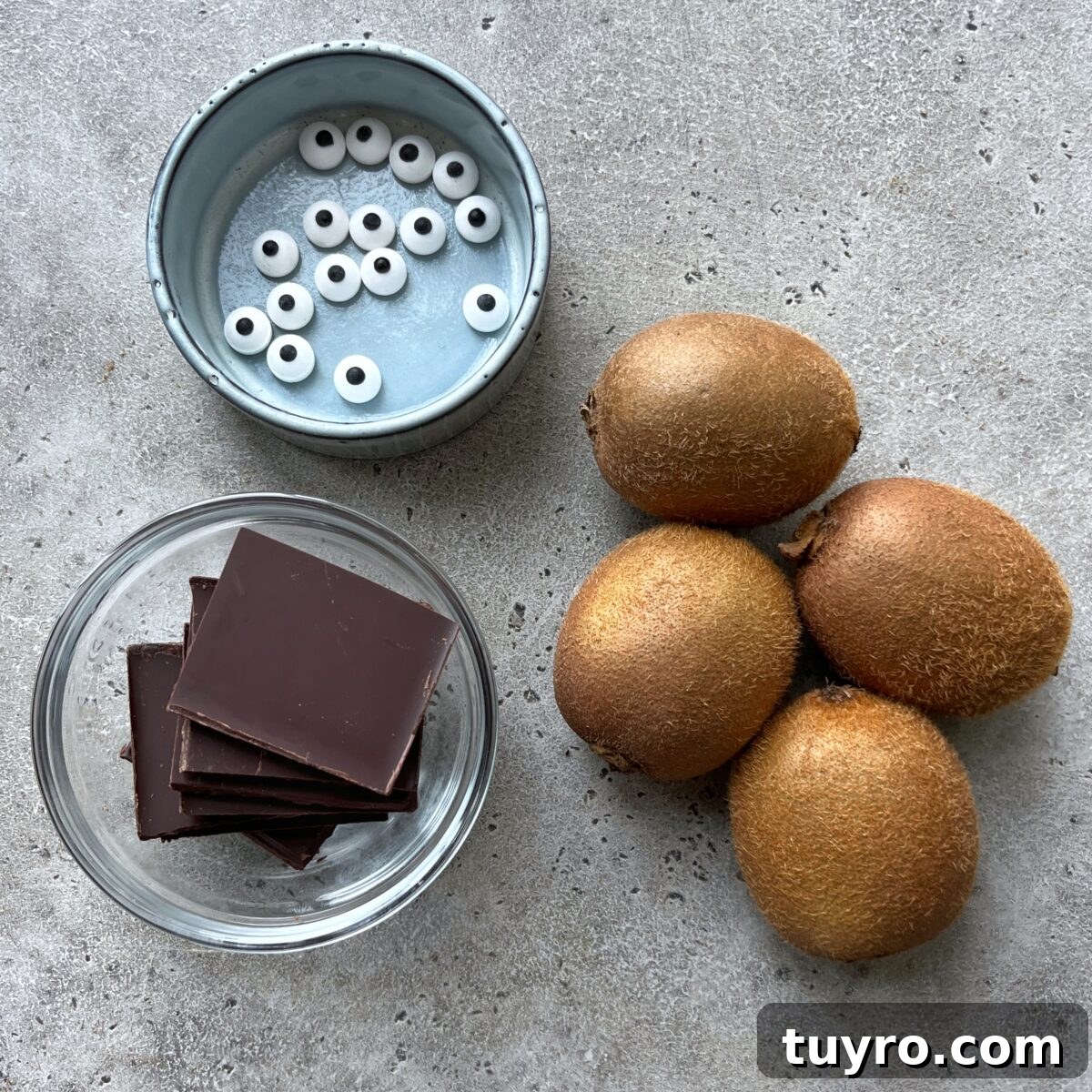 Ingredients needed for Beetlejuice Kiwis featuring a bowl of candy eyeballs, several pieces of dark chocolate in a glass dish, and four whole kiwis.