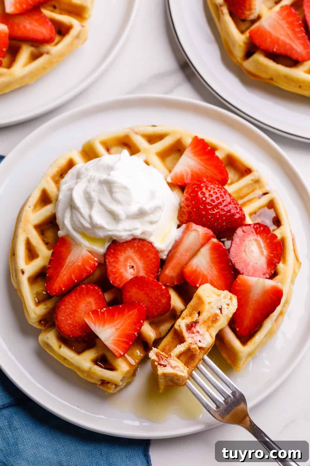 Fluffy Homemade Strawberry Waffles Recipe 6 serving of strawberry waffles topped with whipped cream and fresh cut strawberries and syrup sitting on a white round plate.
