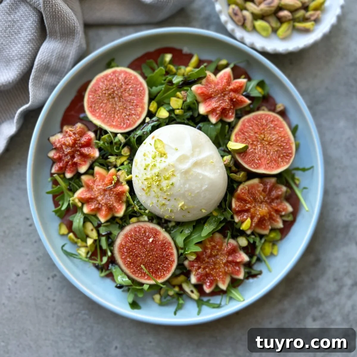 Fig and Bresaola Salad with Arugula, Burrata & Balsamic 3 A plate with burrata cheese and bresaola topped with arugula, surrounded by halved and sliced figs, chopped pistachios, and flower-shaped fig pieces creates a stunning Fig Bresaola Salad. A bowl of pistachios sits nearby.