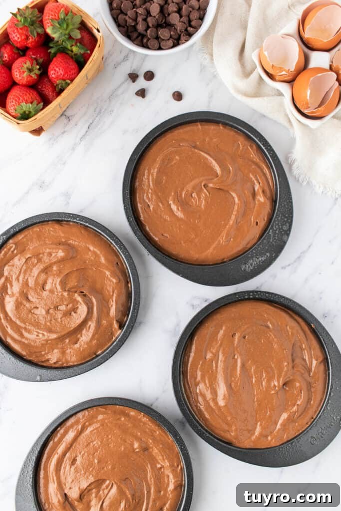Chocolate Strawberry Cake with Rich Ganache 5 Four cake pans filled with chocolate batter, ready for baking, with strawberries and chocolate chips nearby.