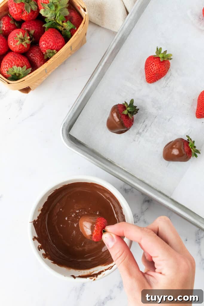 Chocolate Strawberry Cake with Rich Ganache 8 Hand dipping a fresh strawberry into melted chocolate over a parchment-lined baking sheet.