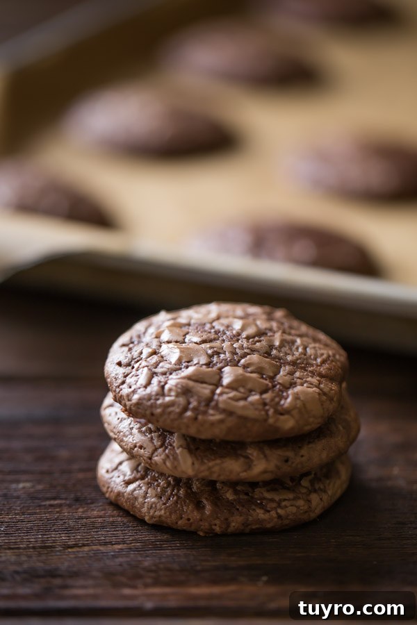 How to Make Soft, Chewy Brownie Cookies 2 A stack of three Soft and Chewy Brownie Cookies, with more cookies on a parchment paper lined baking sheet in the background.