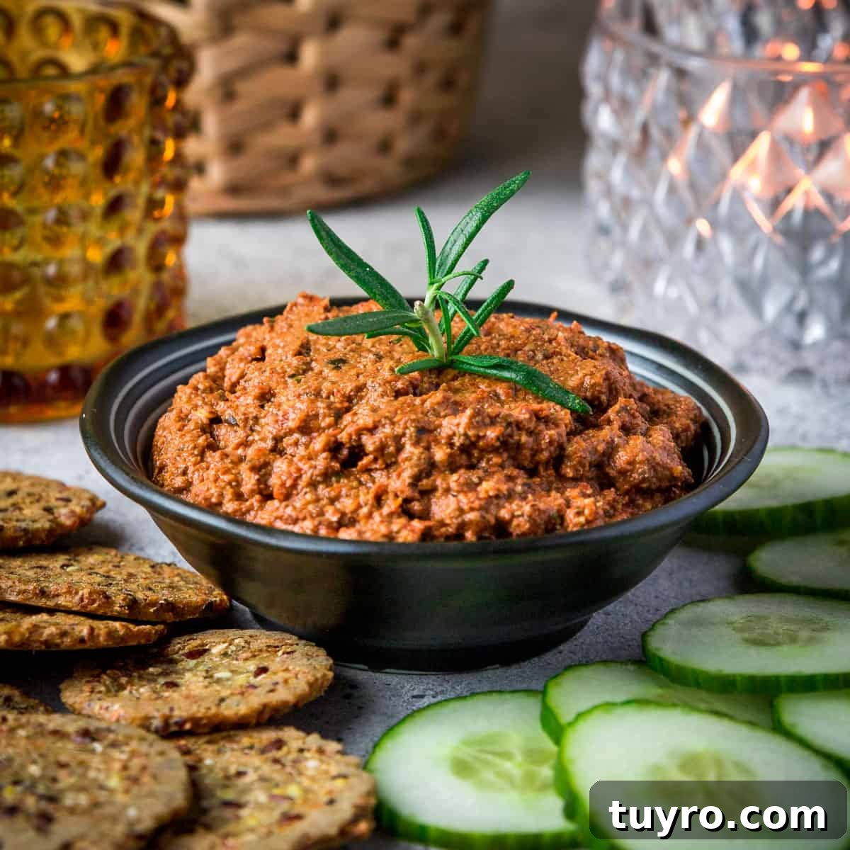 Photo of a party scene with a black bowl full of sundried tomato tapenade garnished with a sprig of rosemary and some crackers and cucumber slices surrounding it.