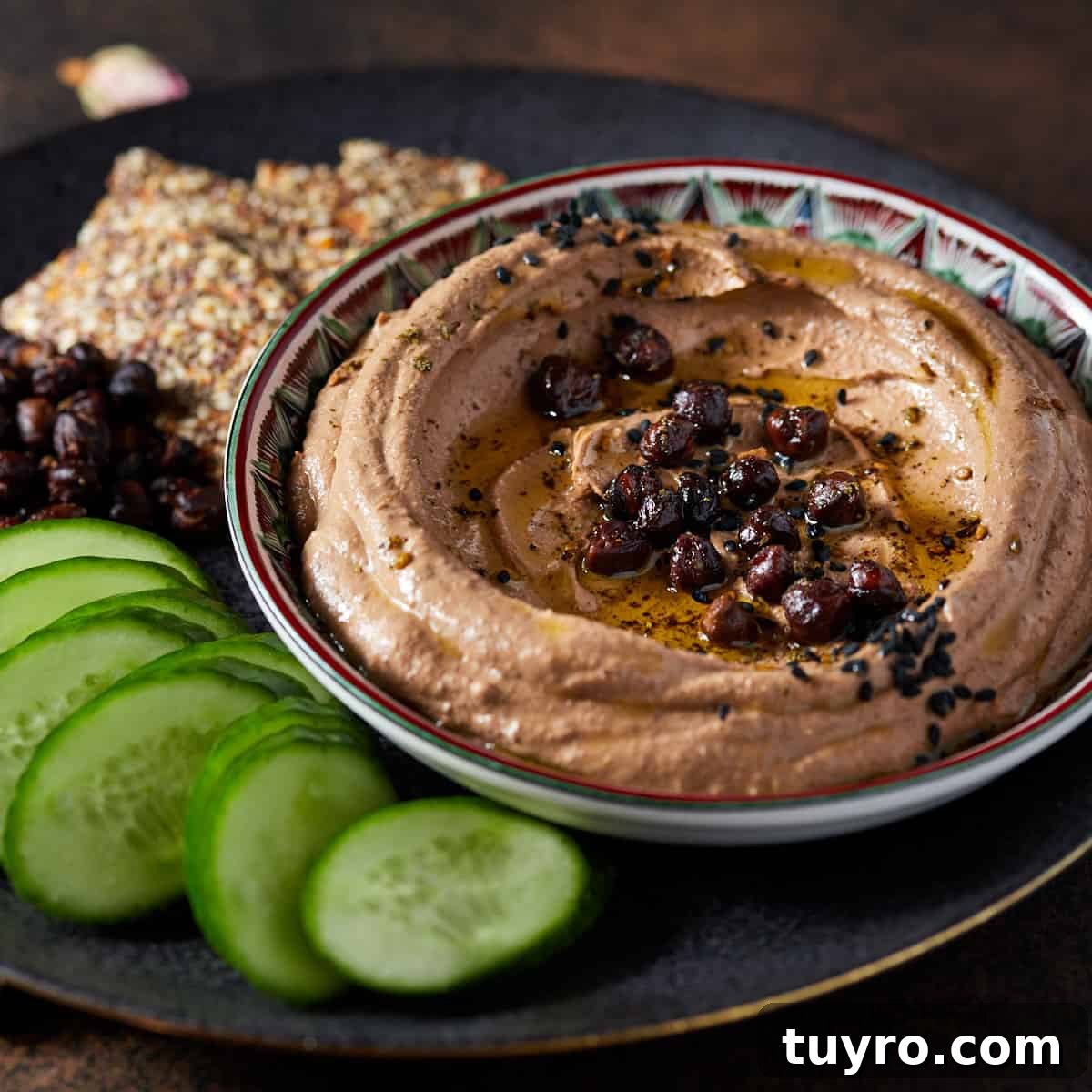 Image of black chickpea hummus garnished and set in a shallow patterned dish, which is set on a black plate with sliced cucumbers, roasted black chickpeas and seeded crackers to the side, all set on a rusty dark background.