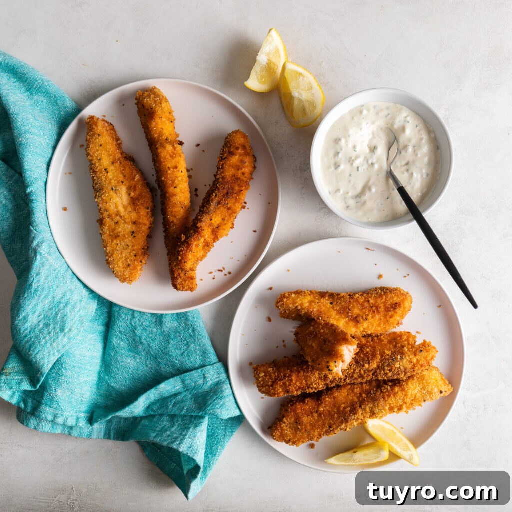 two plates of salmon fish sticks, one with lemon wedges and a bowl of tartar sauce on the side with a spoon in it and a blue dinner napkin
