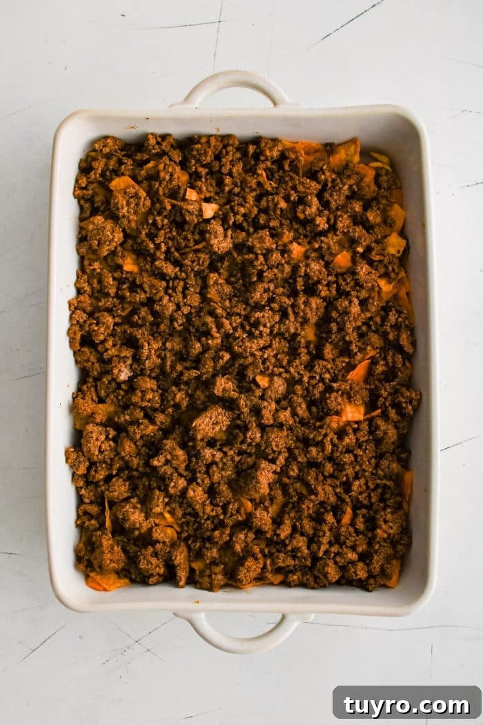Cooked ground beef taco meat layered on top of a double layer of frito and dorito chips in a large white baking dish.