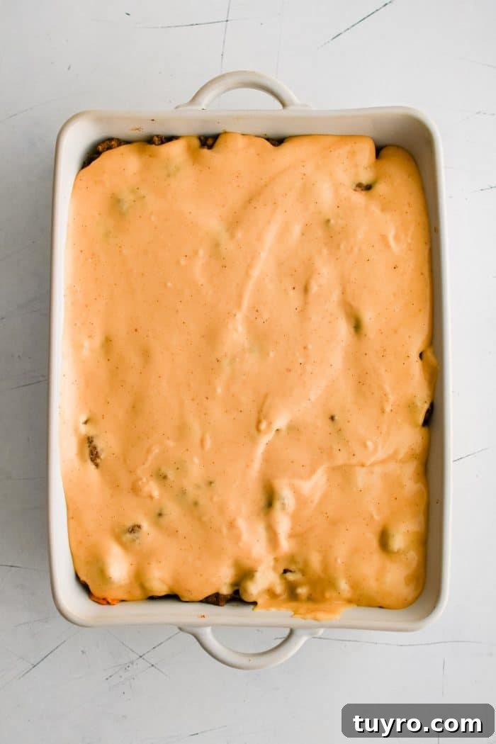 Homemade nacho cheese sauce poured over the ground taco meat layer in a large white baking dish.