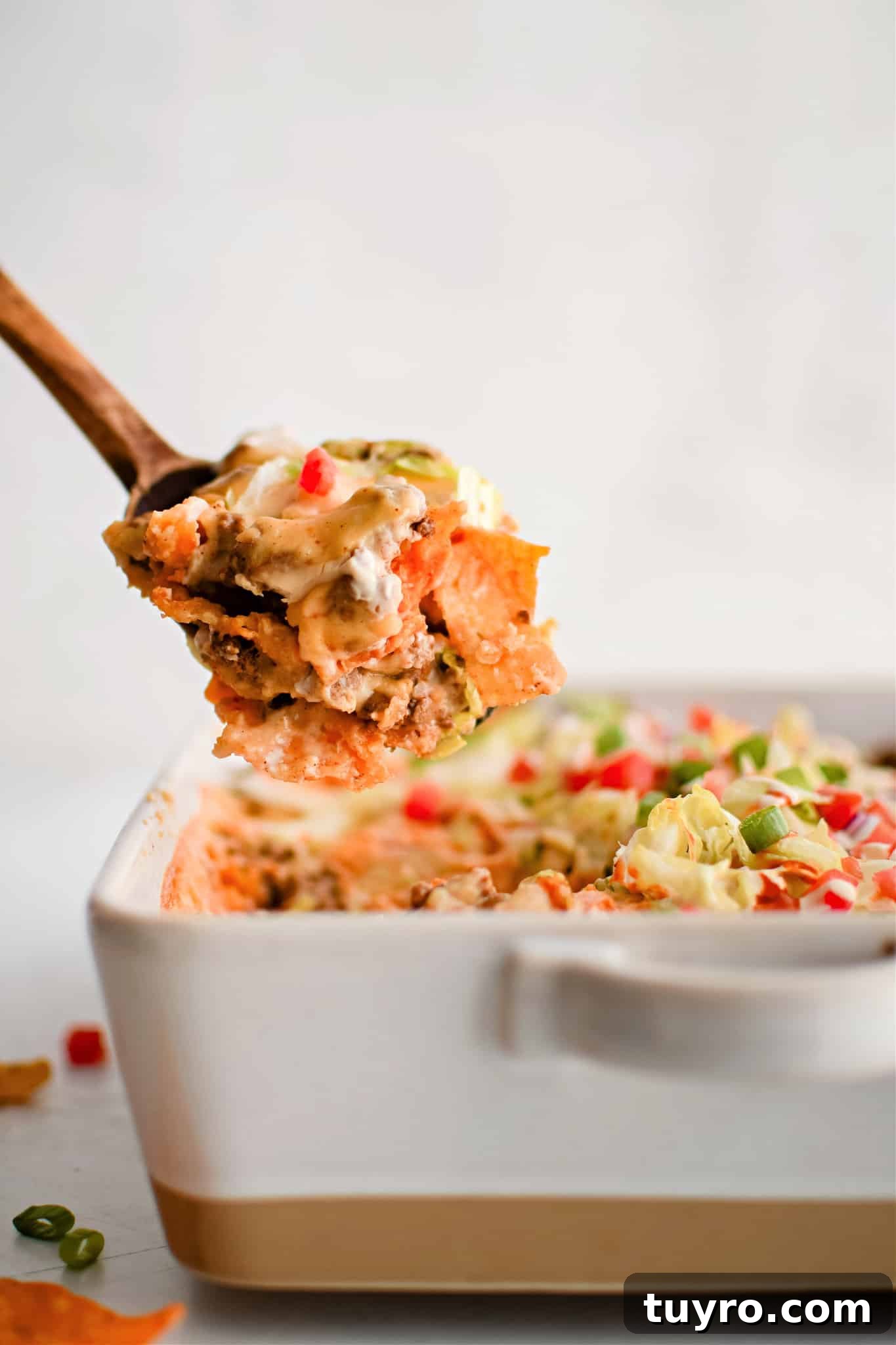 Wooden spatula raising a large portion of walking taco casserole from a white casserole pan filled with the remaining meat and cheese filled casserole.