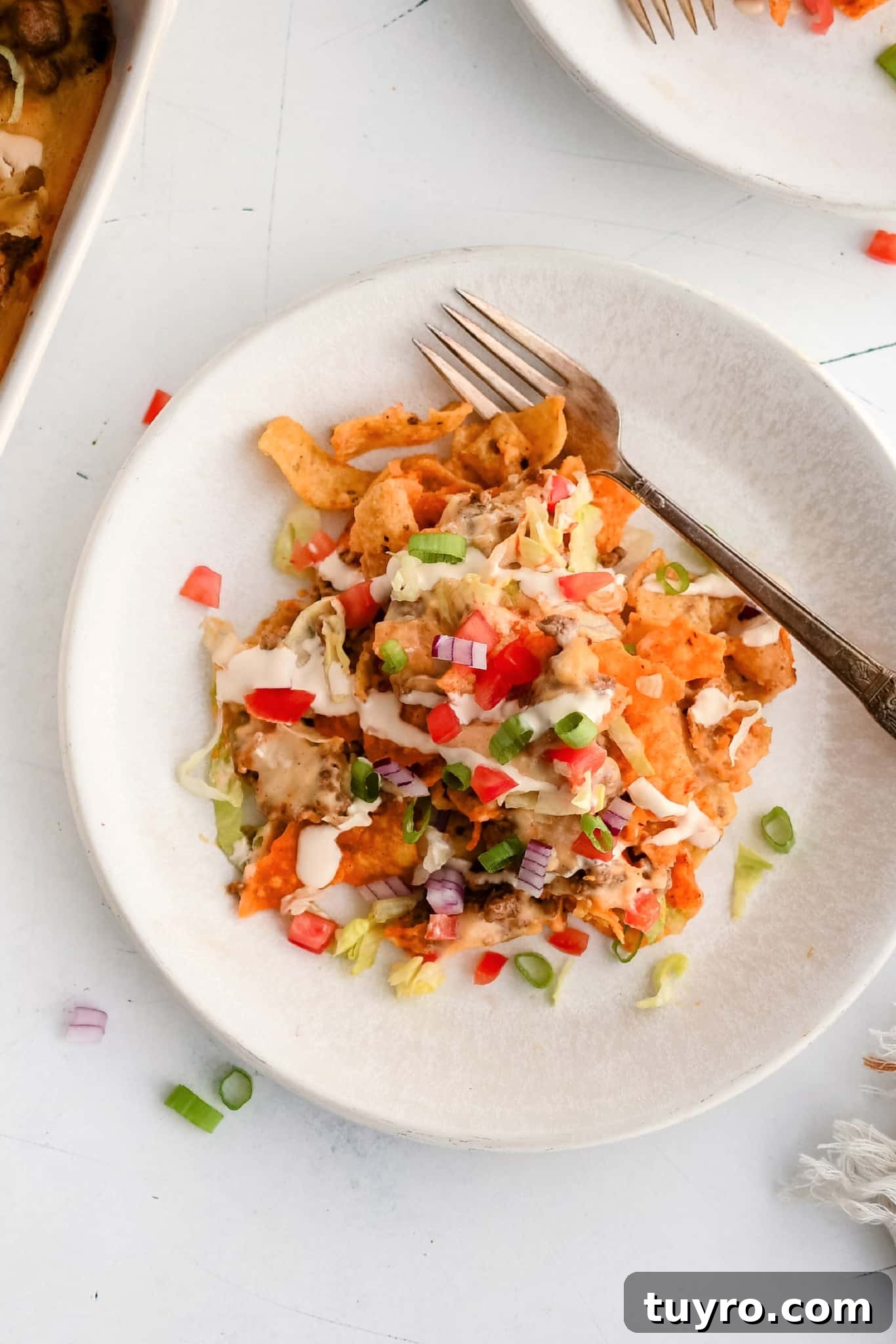 White dinner plate topped with a generous serving of walking taco casserole topped with shredded iceberg lettuce, chopped tomatoes, sour cream, and hot sauce.