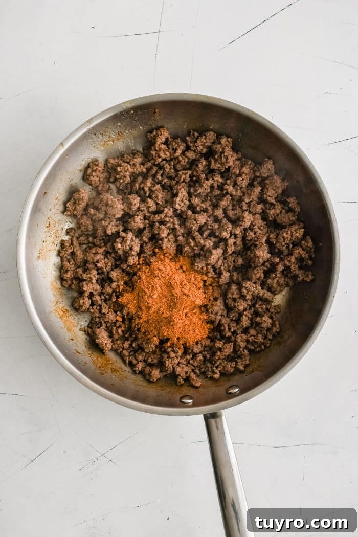 Taco seasoning added to a large stainless steel skillet filled with crumbled browned ground beef.