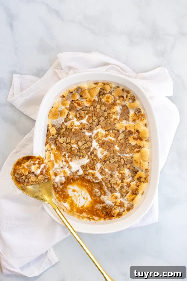 Sweet Potato Casserole with Marshmallows and Oat Streusel in a white baking dish with a gold serving spoon sitting on a white kitchen towel.