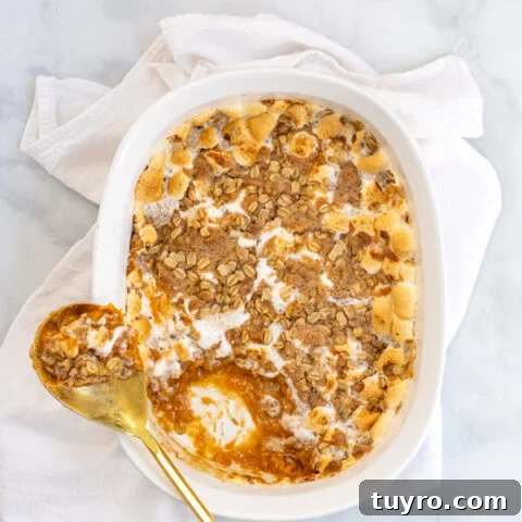 Sweet Potato Casserole with Marshmallows and Oat Streusel in a white baking dish with a gold serving spoon sitting on a white kitchen towel.