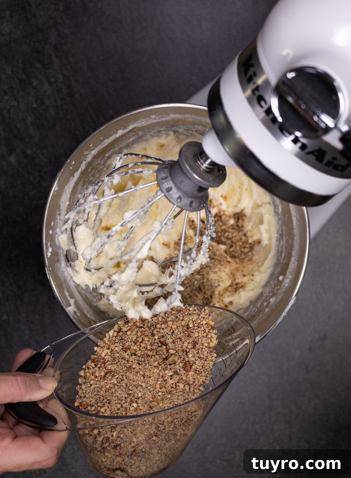 Classic Red Velvet Cake with Cream Cheese Frosting 13 person pouring in chopped pecans into a standing mixer for making cream cheese frosting.