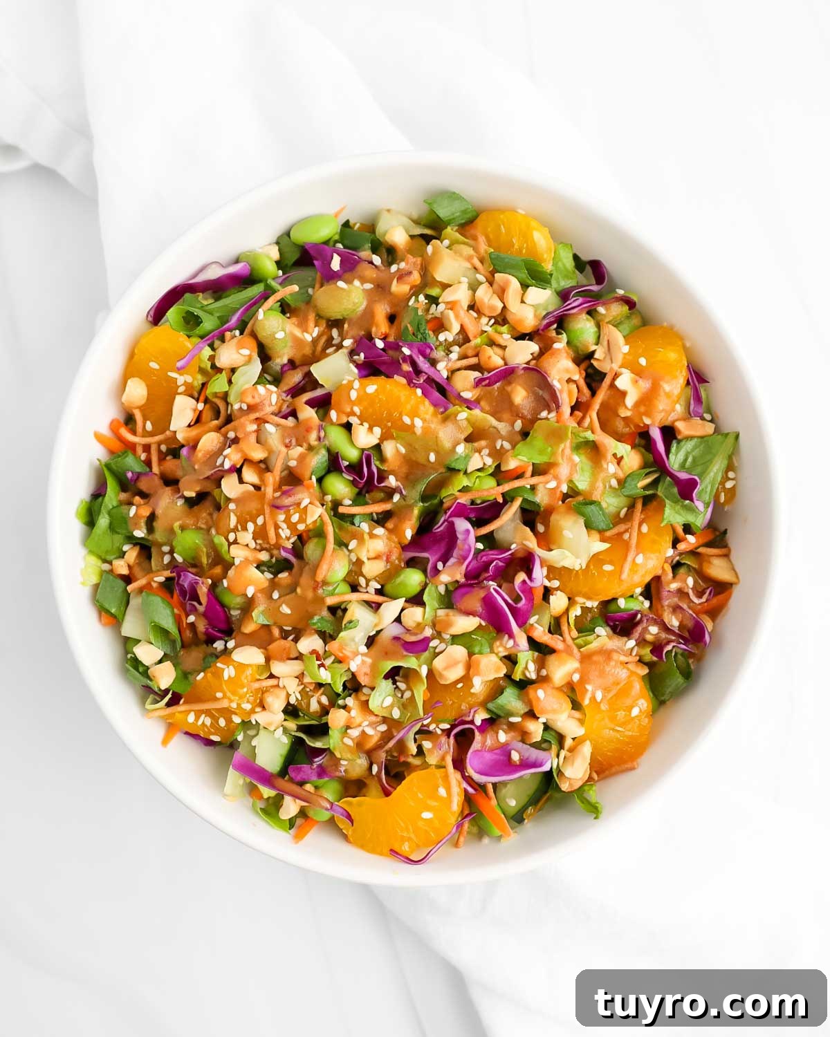 A white bowl filled with a fresh salad made with lettuce, cabbage, oranges, chow mein noodles, edamame, and a peanut sauce.