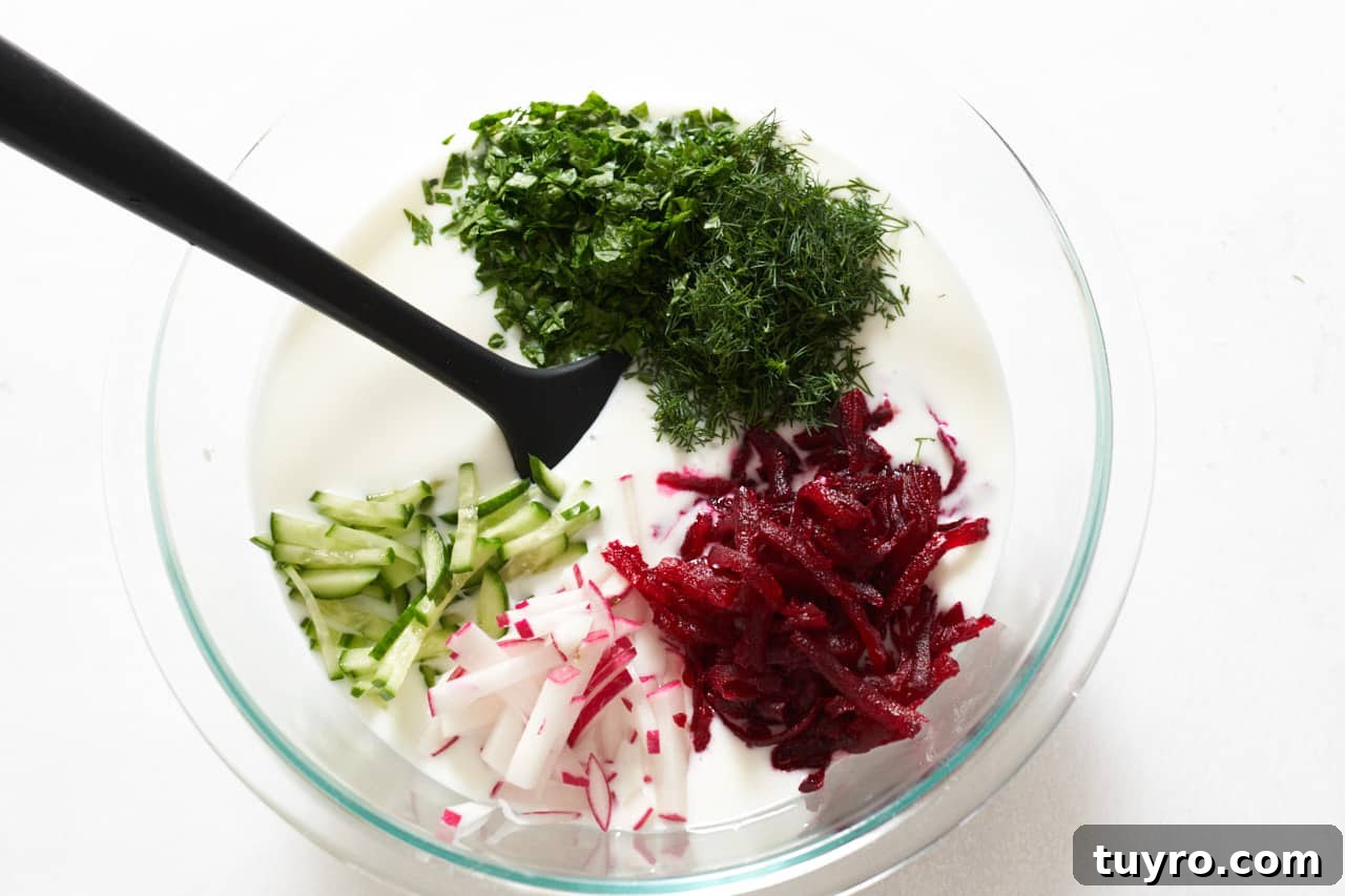 Chilled Beet Borscht: Classic Cold Soup Recipe 7 Chopped dill and parsley, and sliced beets, cucumber, and radishes being added to a bowl of kefir with a black spatula in it.