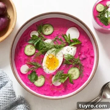 Chilled Beet Borscht: Classic Cold Soup Recipe 9 A bowl and a small glass of cold borscht with toppings including sliced cucumbers and radishes, fresh herbs, and a hard boiled egg. Fresh herbs, two spoons, a blue napkin, and a bowl of radishes surround the soups.